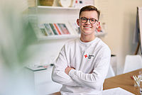 Ein junger Mann in einem weißen Pullover mit Logo des DRK auf der Brust. Er lacht in die Kamera und hat die Arme vor der Brust verschränkt.