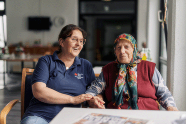 Zwei Frauen sitzen nebeneinander und unterhalten sich. Die linke Frau arbeitet beim DRK, trägt ein dunkelblaues DRK Shirt und hat dunkle Haare. Die rechte Frau ist etwas älter als die linke und trägt ein Kopftuch.