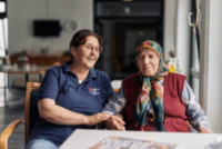 Zwei Frauen sitzen nebeneinander und unterhalten sich. Die linke Frau arbeitet beim DRK, trägt ein dunkelblaues DRK Shirt und hat dunkle Haare. Die rechte Frau ist etwas älter als die linke und trägt ein Kopftuch.